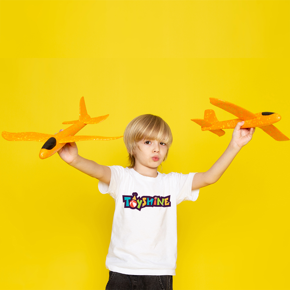 Child in a white Toyshine t-shirt holding two orange toy aeroplanes against a yellow background